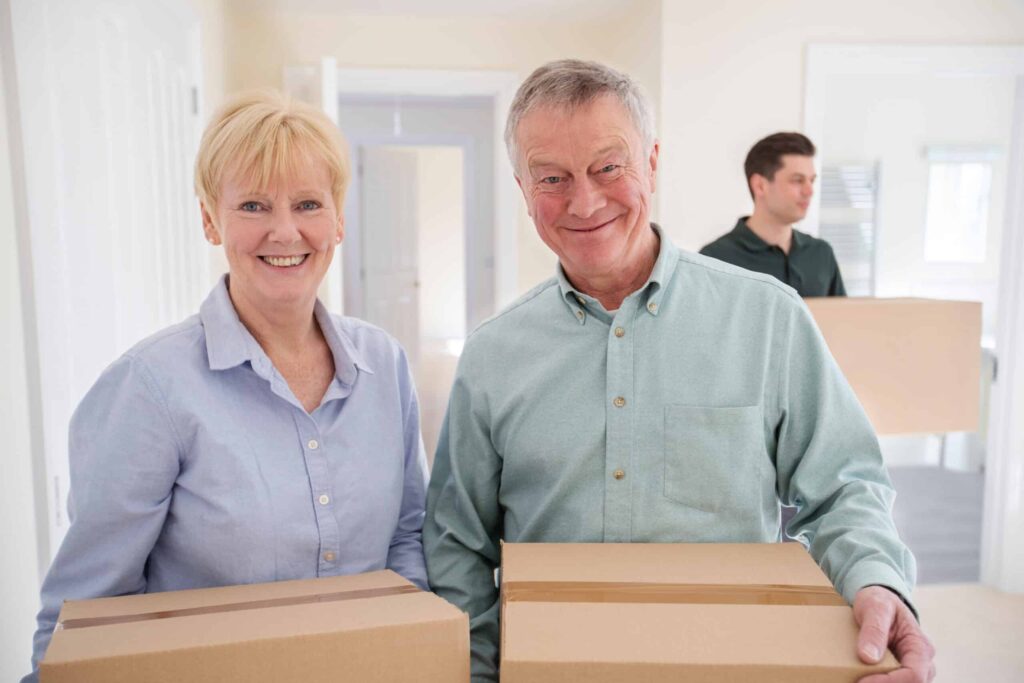 Two older adults smiling while holding cardboard boxes indoors, with a younger person carrying a box in the background.