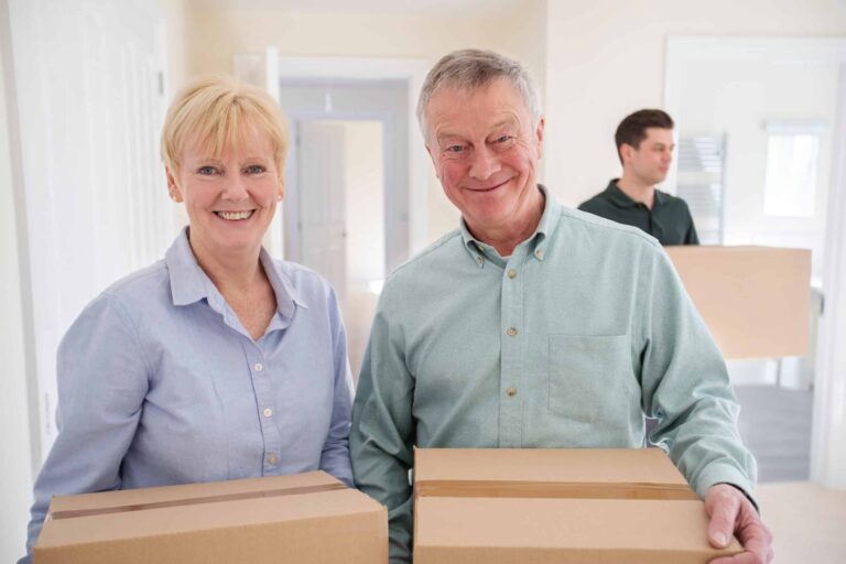 Two older adults smiling while holding cardboard boxes indoors, with a younger person carrying a box in the background.