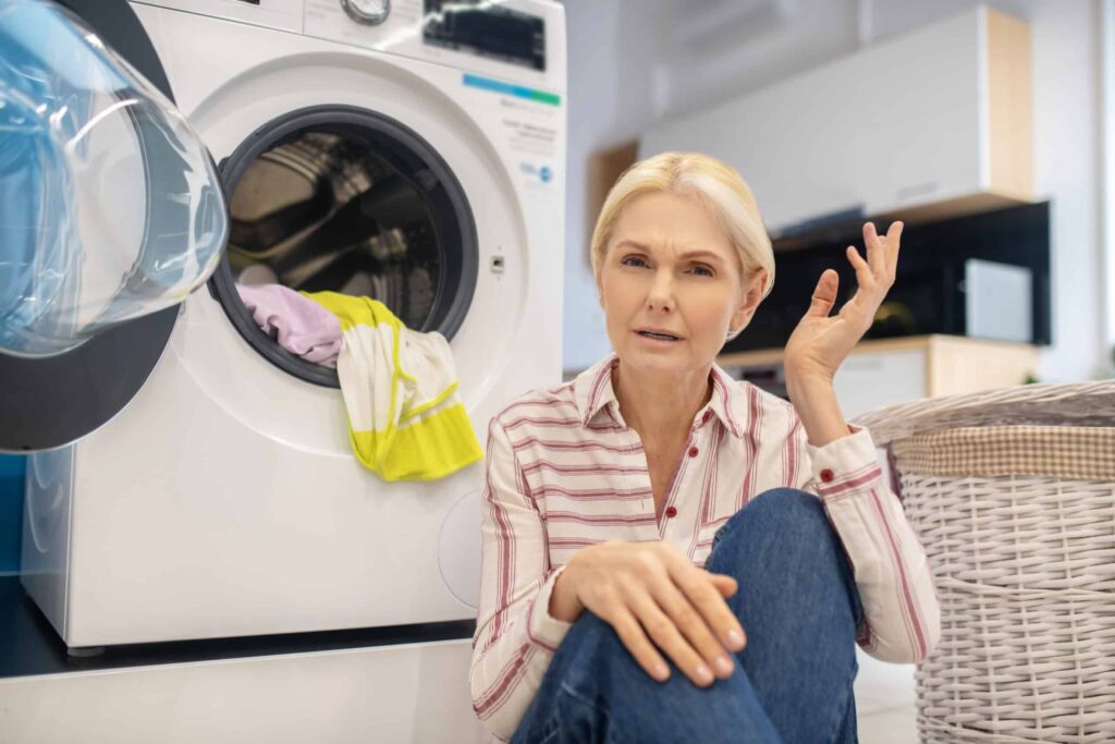 A woman sits on the floor in front of a washing machine with clothes inside. She has one hand raised and an expression suggesting confusion or frustration. A laundry basket is nearby.