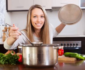 Woman smiling in kitchen, lifting lid from a pot on the stove, holding a ladle. Vegetables and herbs are on the countertop.