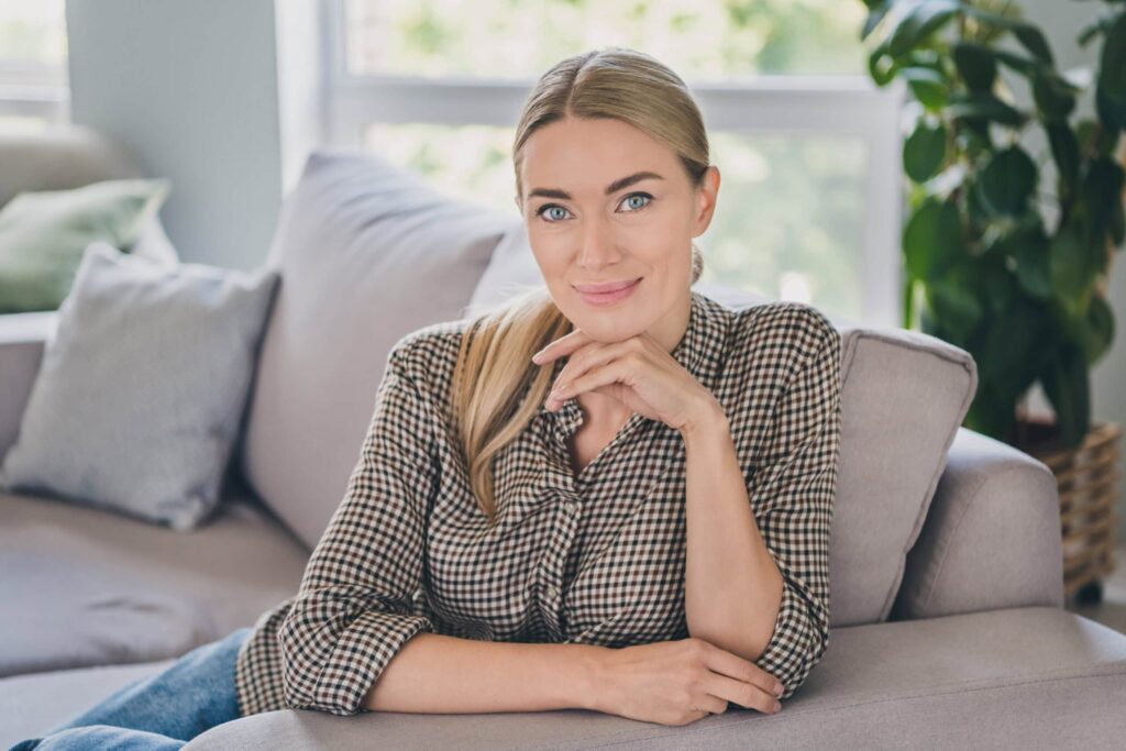 A person with long hair, wearing a checkered shirt, sits on a couch, resting their chin on their hand. There are cushions and a potted plant in the background.