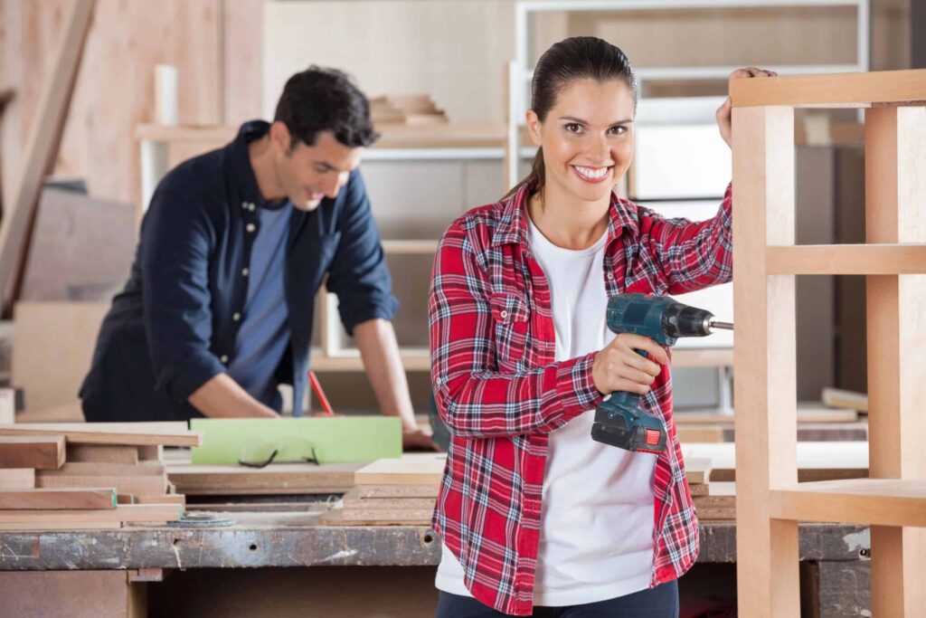 A woman in a plaid shirt holds a power drill, smiling at the camera. A man in the background works on a woodworking project.