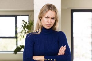 Person with long hair in a blue sweater stands with arms crossed, displaying a serious expression, in front of a neutral background with windows.