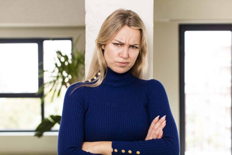 Person with long hair in a blue sweater stands with arms crossed, displaying a serious expression, in front of a neutral background with windows.