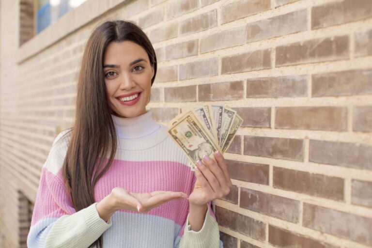 A woman with long hair smiles while holding a fan of U.S. dollar bills against a brick wall.
