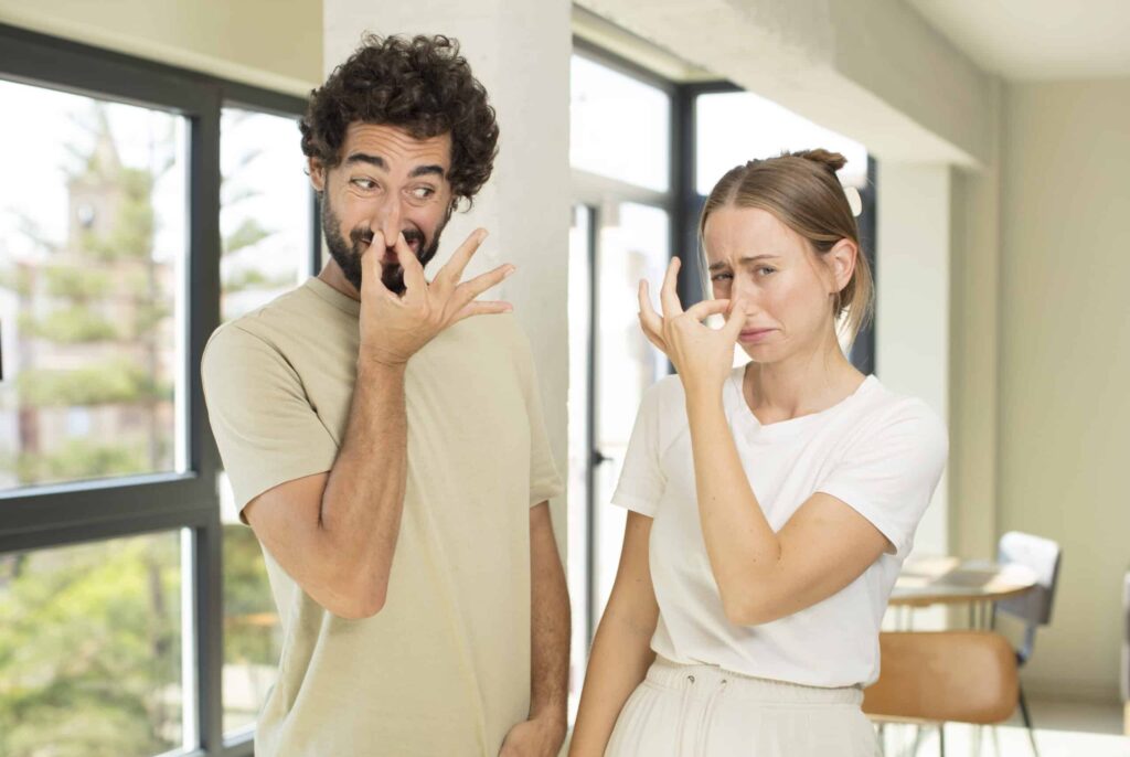 Two people stand indoors, pinching their noses and making faces as if reacting to an unpleasant smell.