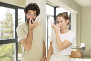 Two people stand indoors, pinching their noses and making faces as if reacting to an unpleasant smell.
