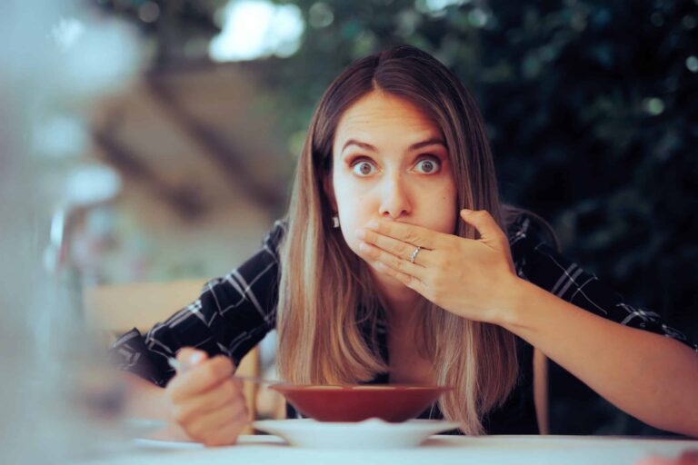 A woman sitting at a table covers her mouth with one hand while holding a spoon with the other. A bowl of soup is in front of her.