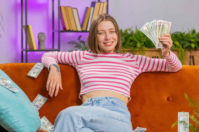 A person sits on an orange couch, holding a fan of US dollar bills with more money scattered around. They are wearing a pink striped shirt and jeans, smiling at the camera. Books are on a shelf in the background.