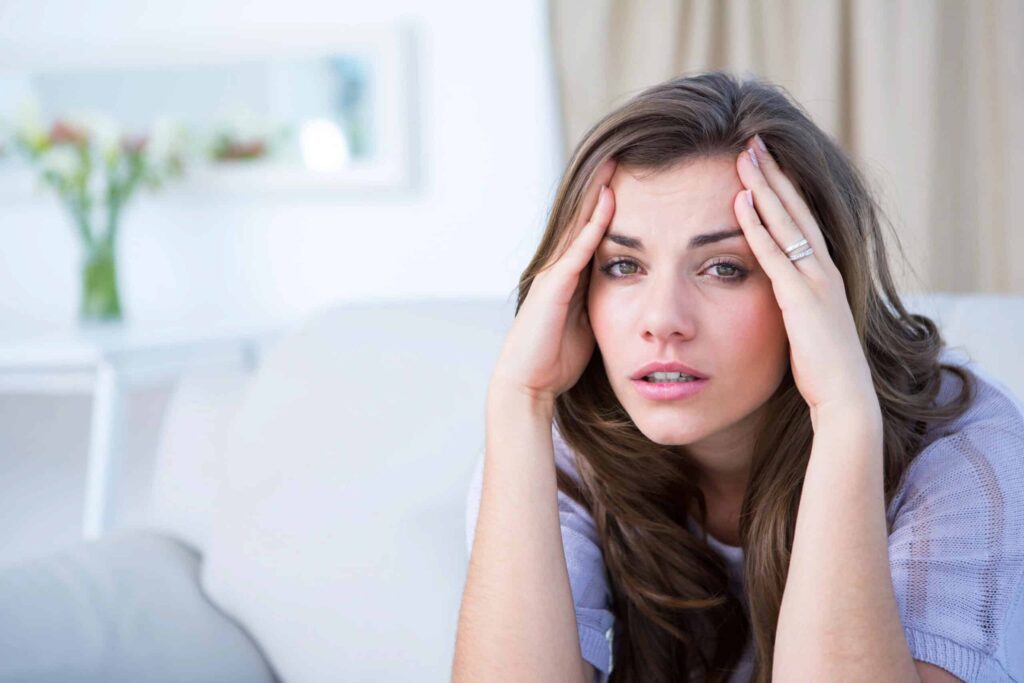 A woman sitting on a couch holds her head with both hands, looking distressed. A vase of flowers and a mirror are in the blurred background.
