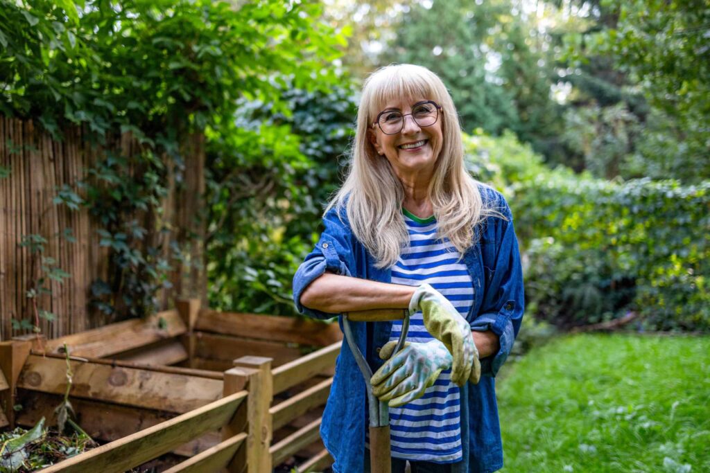 A woman with long hair and glasses smiles while standing in a garden, wearing gloves and holding a gardening tool. There is foliage and wooden fencing in the background.