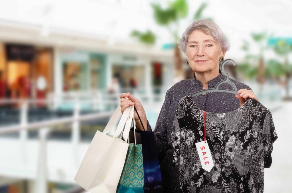 An older woman holds several shopping bags and a dress on a hanger with a sale tag in a mall setting.