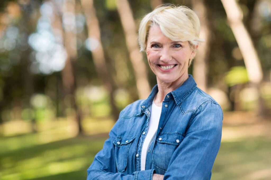 A smiling woman with short blonde hair stands outdoors with her arms crossed, wearing a blue denim shirt and a white top. Trees and greenery are blurred in the background.