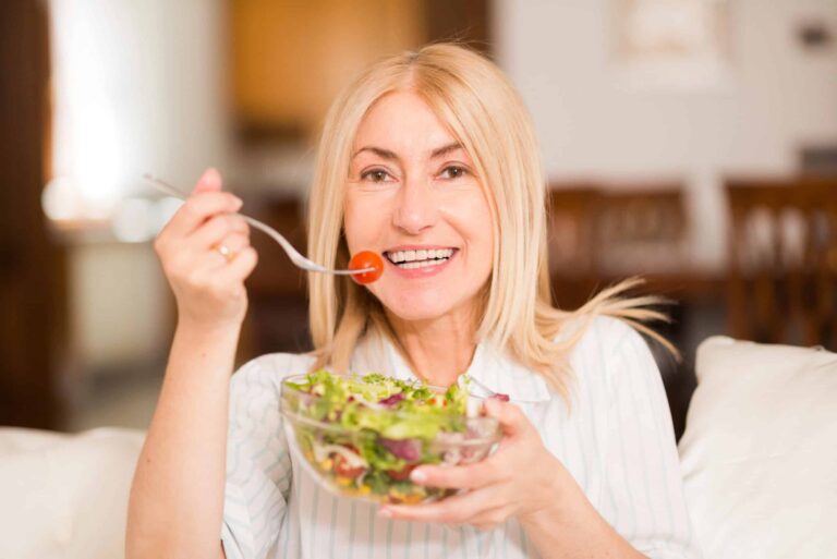 A woman with blonde hair smiles while holding a fork with a cherry tomato and a bowl of salad.