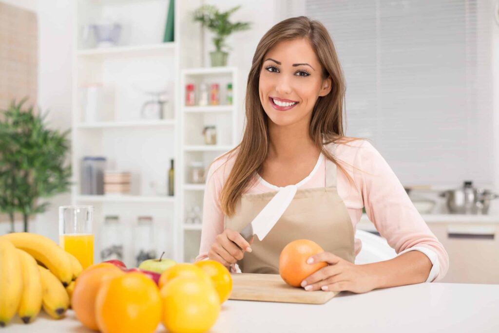 Woman in an apron smiling and holding a knife while cutting fruit in a kitchen. Nearby are bananas, apples, oranges, and a glass of orange juice.