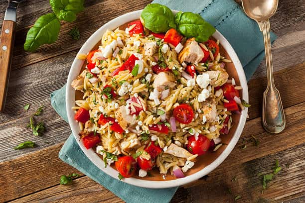A bowl of orzo pasta salad with grilled chicken, cherry tomatoes, feta cheese, red onions, and herbs, garnished with basil.