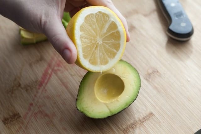 A hand holding a halved lemon above a halved avocado on a wooden cutting board, with a knife in the background.