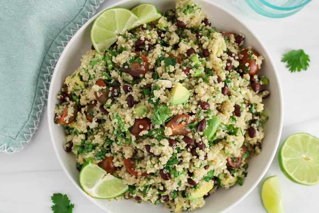 A bowl of quinoa salad with black beans, cherry tomatoes, avocado, cilantro, and lime wedges.