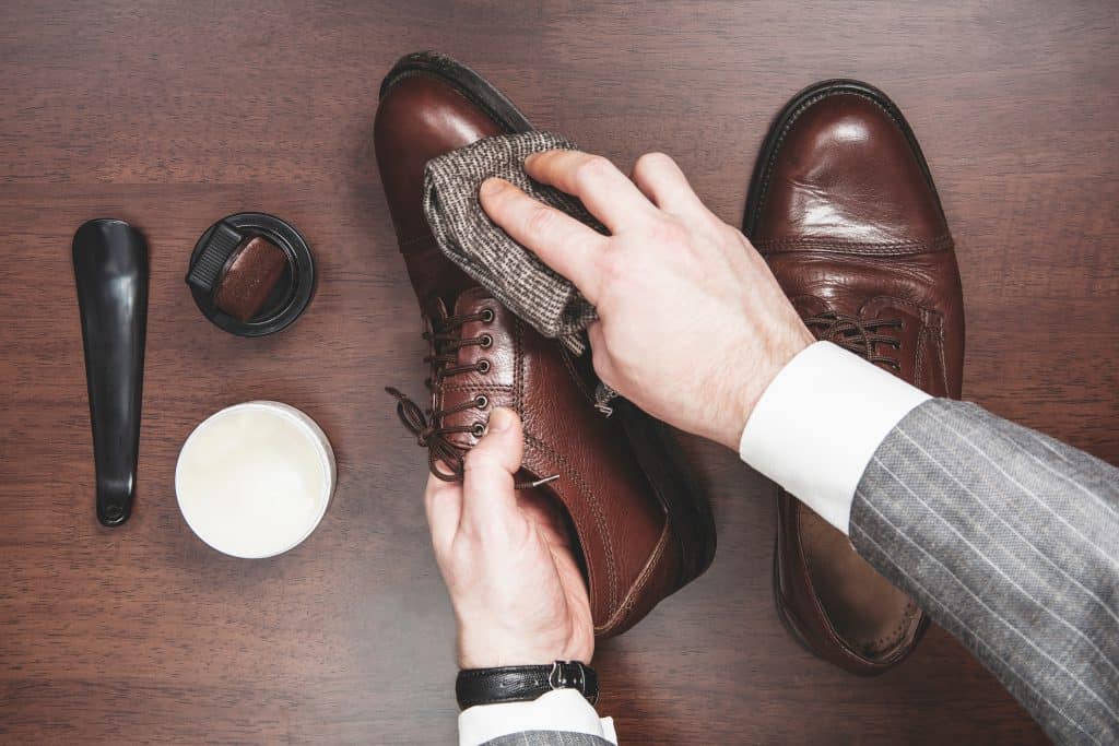 A person in a suit polishes brown leather shoes with a cloth on a wooden surface. Nearby are a shoe brush, polish, and cream.