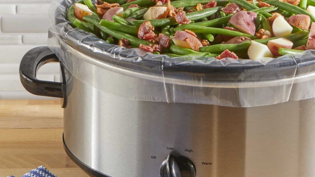 A slow cooker filled with green beans, chopped potatoes, and bacon pieces, lined with a clear plastic liner.