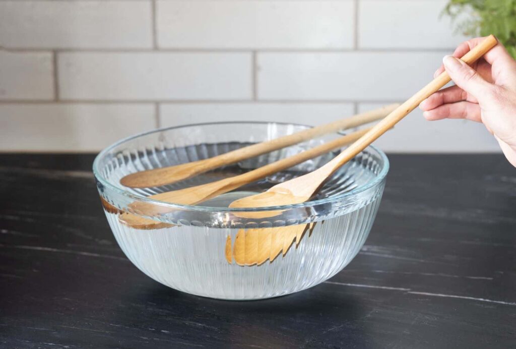 A hand holding wooden spoons above a glass bowl filled with water on a dark countertop.
