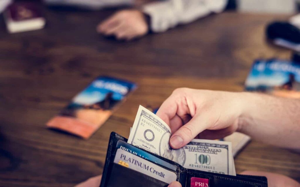 A person holding a wallet containing a credit card is pulling out a one-hundred-dollar bill.