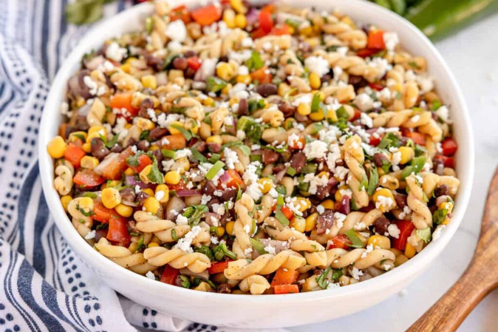 A white bowl of pasta salad with fusilli, corn, black beans, red bell peppers, and feta cheese on a striped cloth.