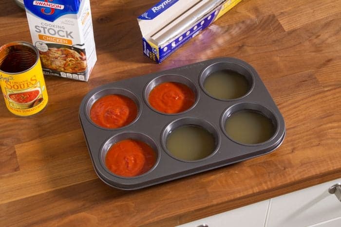 A muffin tray on a wooden counter, with some cups filled with red tomato soup and others with chicken stock. Nearby are soup and stock containers and a roll of aluminum foil.