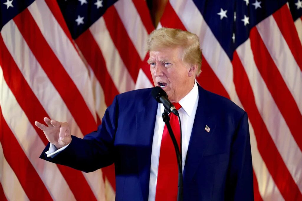 Trump in a suit gestures while speaking at a podium with multiple American flags in the background.