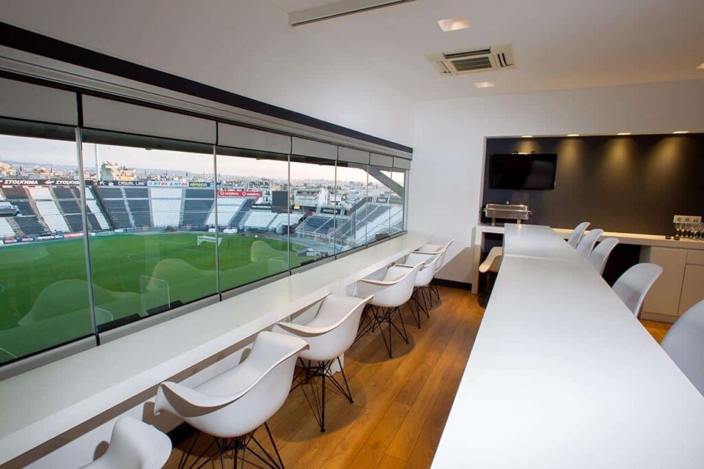 Stadium viewing suite with modern chairs, long tables, and a large glass window overlooking the field.