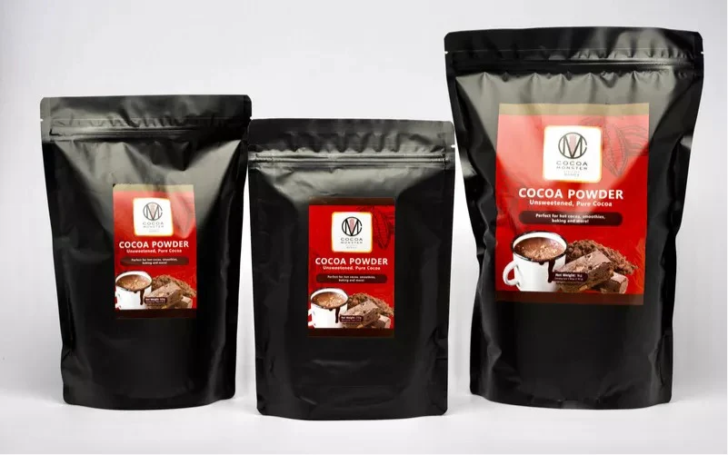 Three black packages of cocoa powder with red labels and images of a cup of cocoa and chocolate pieces are displayed. The packages have varied sizes and the brand logo is visible.