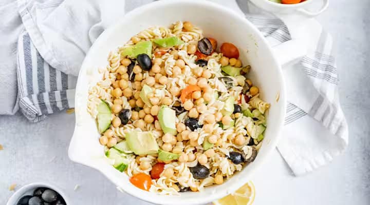 A bowl of pasta salad with chickpeas, avocado, cherry tomatoes, black olives, and zucchini, placed on a white surface with a striped cloth nearby.
