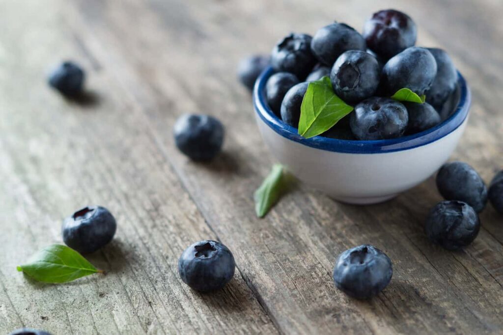 A small white bowl filled with blueberries and a few green leaves sits on a wooden surface, surrounded by scattered blueberries.