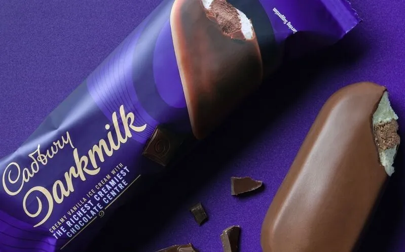 A Cadbury Darkmilk ice cream bar is unwrapped on a purple surface, with chocolate pieces scattered around.