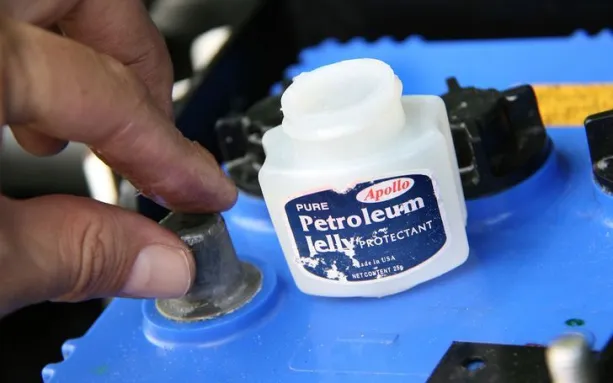Hand applying petroleum jelly on a battery terminal with an open container of petroleum jelly nearby.