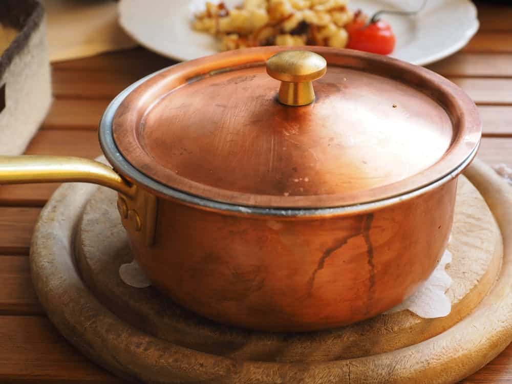 A copper pot with a brass handle and lid sits on a wooden surface. A plate with food is blurred in the background.