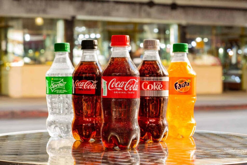 Five soft drink bottles on a table, featuring Sprite, Coca-Cola, Diet Coke, and Fanta against a blurred urban background.