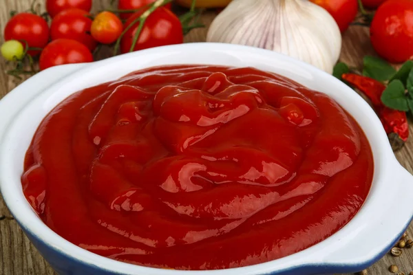 A bowl of tomato ketchup surrounded by fresh tomatoes and garlic on a wooden surface.