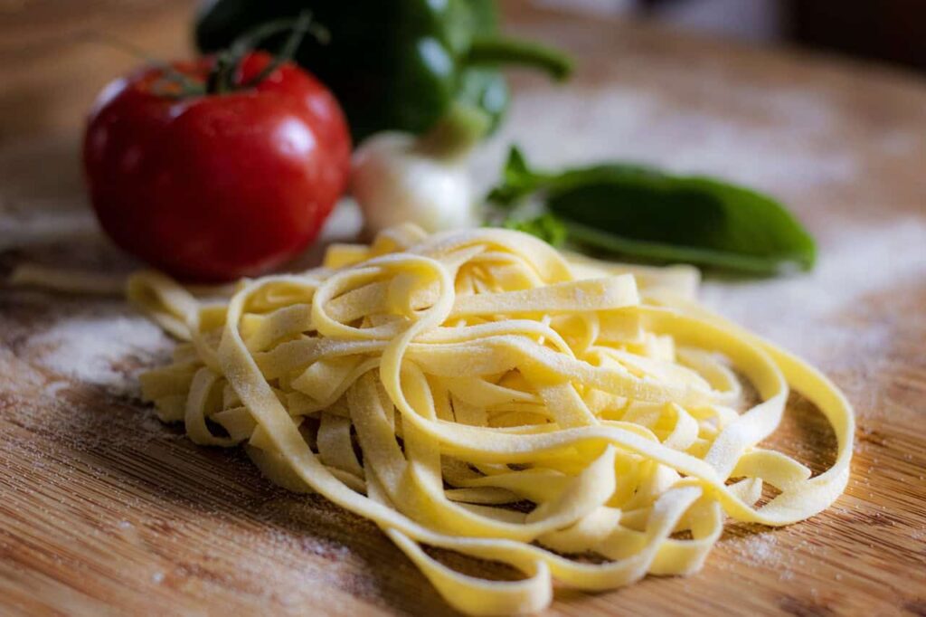 A pile of uncooked pasta on a wooden surface with a tomato, green bell pepper, garlic, and basil leaves in the background.