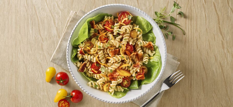 A bowl of pasta salad with tomatoes, peppers, and bacon on lettuce leaves. Fork and knife beside the bowl.