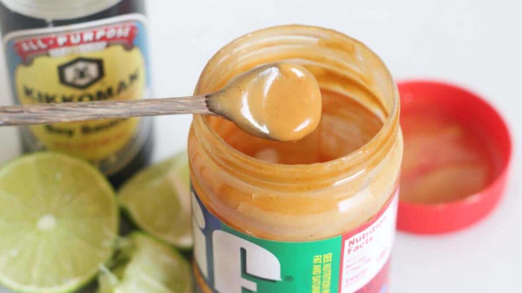 Open jar of peanut butter with a spoon, surrounded by lime halves and a bottle of soy sauce.