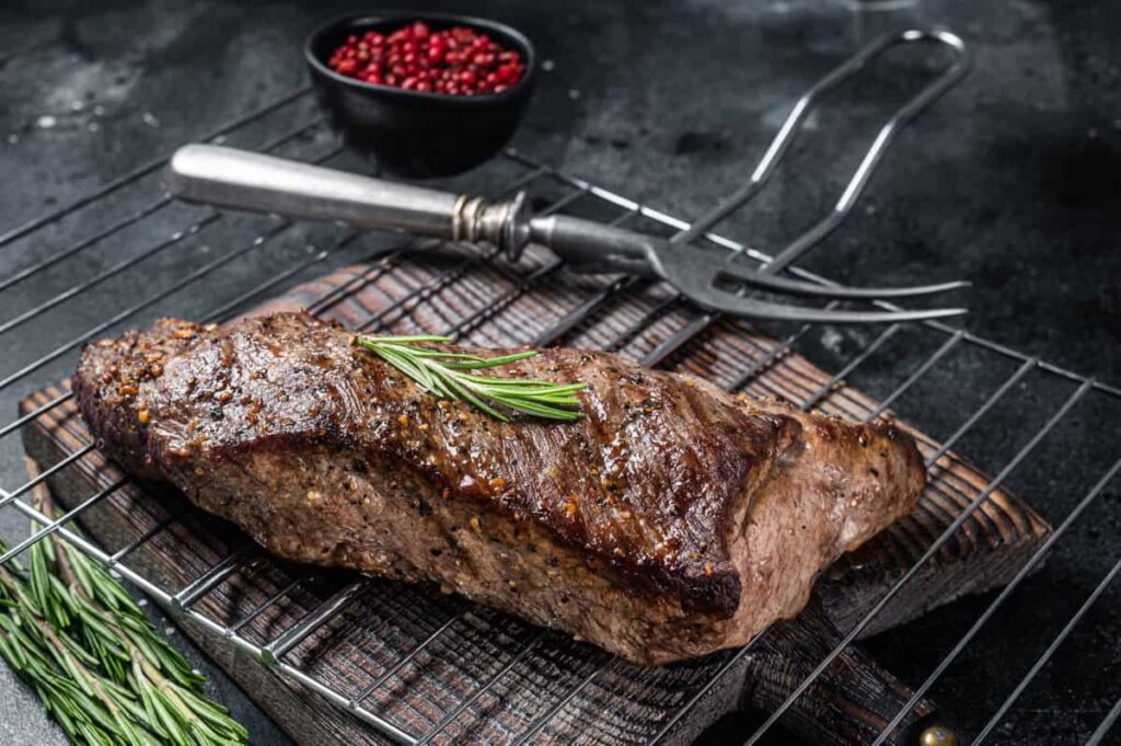 Grilled steak with rosemary garnish on a wooden board, accompanied by a fork and a bowl of red peppercorns.