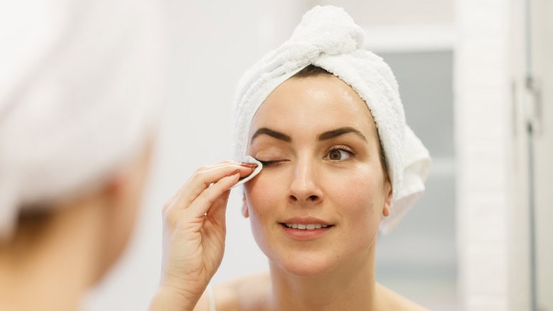 Woman with a towel on her head applies makeup remover to her eye with a cotton pad in front of a mirror.