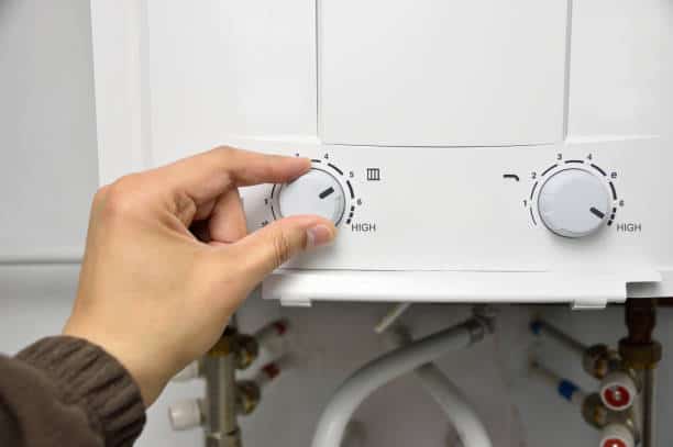 Hand adjusting a temperature dial on a white gas water heater with visible pipes below.