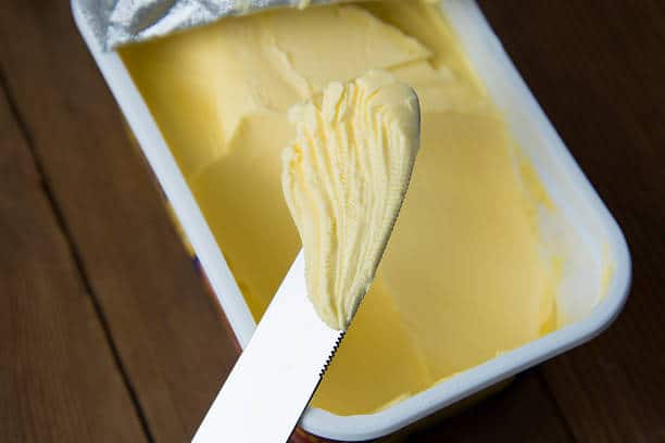 A knife holds a portion of margarine over an open rectangular container of margarine on a wooden surface.
