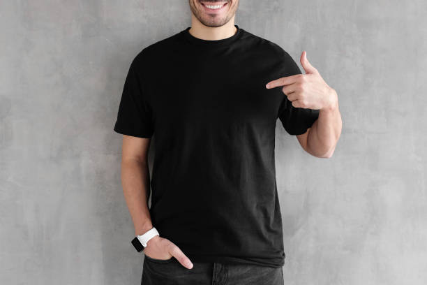 Person wearing a black t-shirt and a smartwatch smiles while pointing to their shirt against a gray background.