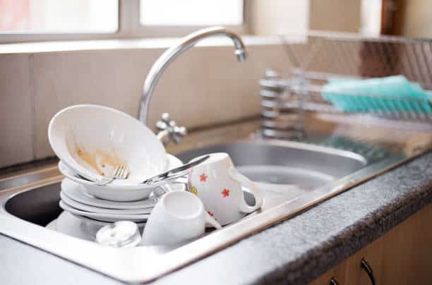 A kitchen sink filled with dirty dishes, including plates, cups, and cutlery, beside a clean dish rack.