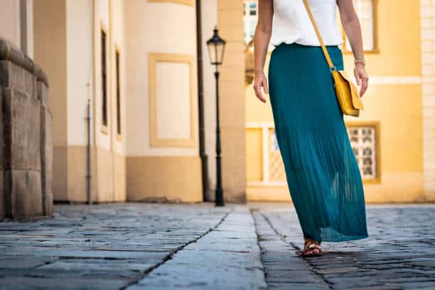A person in a white top and teal skirt walks on a cobblestone street, with a yellow purse over their shoulder. Buildings and a lamp post are in the background.