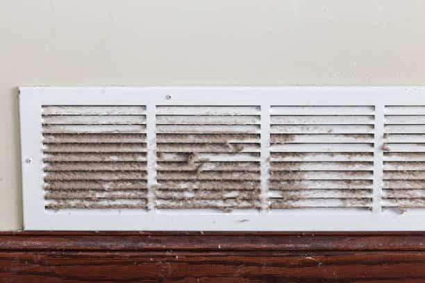 A dirty air vent covered in dust and debris is installed on a wall above a wooden baseboard.