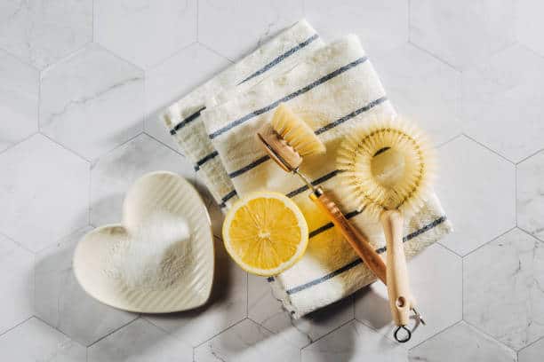 Wooden scrub brushes, a half lemon, a heart-shaped dish with white powder, and a striped towel on a hexagonal marble surface.